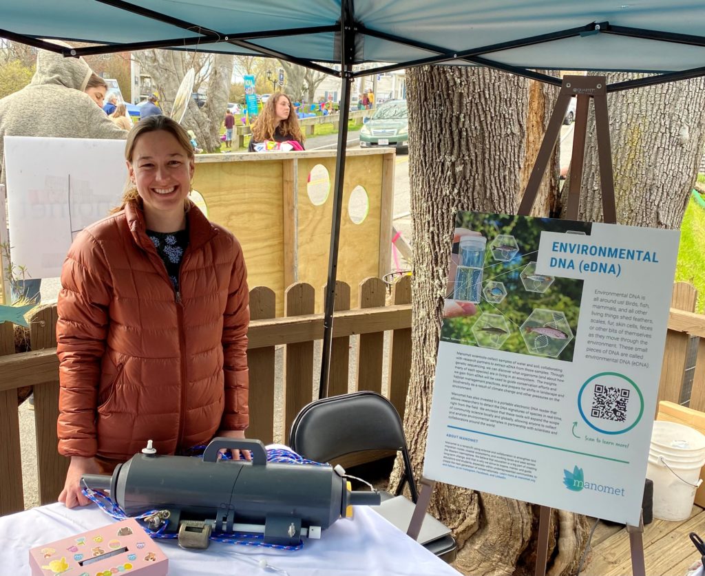 A woman in a rust-orange jacket smiles and stands under a tent next to an informational poster about eDNA