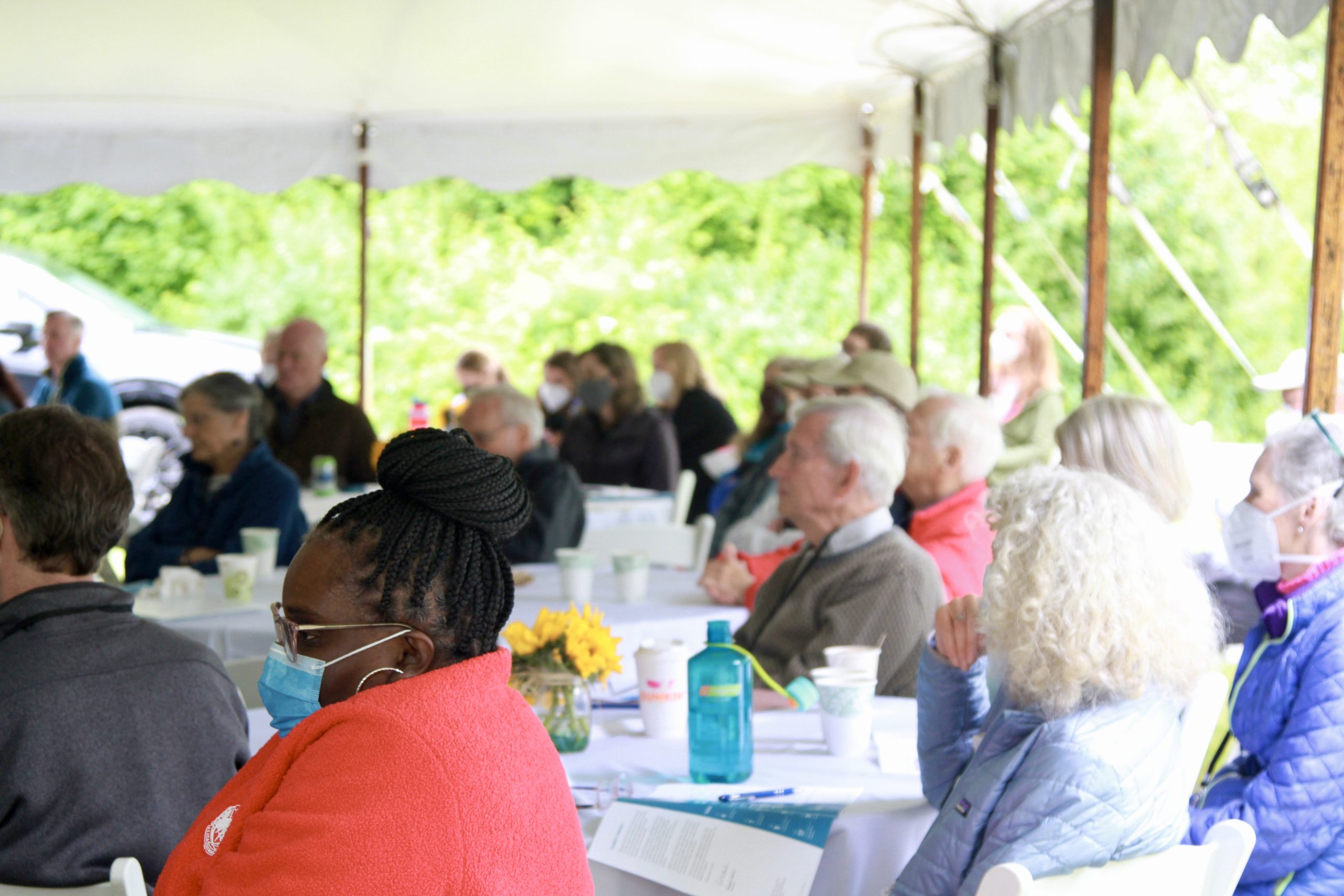 Crowd of people seated under a tent listen intently to a piece of music played over a loudspeaker.